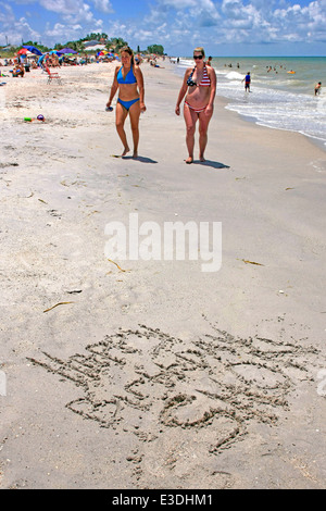 Eine schwangere Frau und ihr Freund zu Fuß am Strand in Florida, wo jemand einen Geburtstagsgruß geschrieben hat Stockfoto