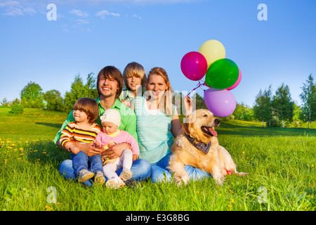 Lächelnde Familie sitzen auf dem Rasen mit Luftballons und Hund Stockfoto