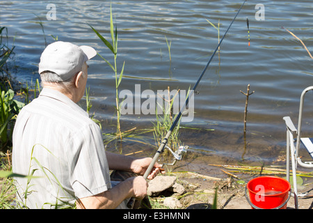 Älterer Mann genießen eine Tage Angeln auf einer Lagune oder See sitzen mit dem Rücken zur Kamera hält eine Rute und Rolle Stockfoto