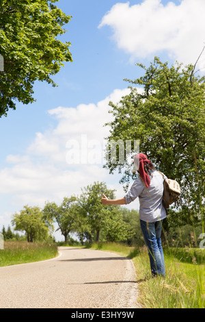 Rückansicht des eine Anhalterin auf einer kleinen Straße, die Daumen hochhalten Stockfoto