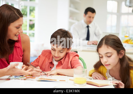 Mama hilft Kindern bei den Hausaufgaben, wie Papa im Hintergrund arbeitet Stockfoto