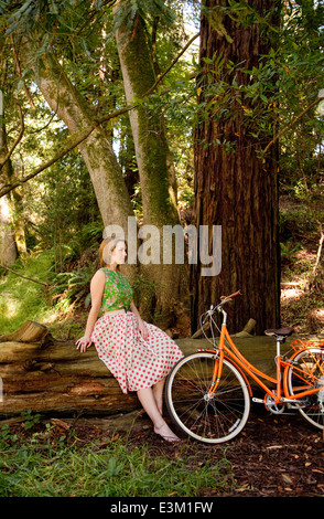 Frau sitzt auf Log in Wald, stützte sich neben ihr Fahrrad Stockfoto