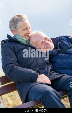 gerne älteres Paar gemeinsam entspannen in der Sonne auf einer Holzbank in der Landschaft mit einer stützenden voller Länge auf Stockfoto