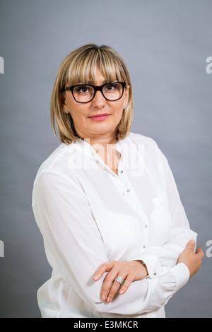 Wissenschaftliche attraktiven Frau mittleren Alters blond in Gläsern Stand mit verschränkten Armen gegen eine graue Zeitmessung in die Kamera schaut Stockfoto