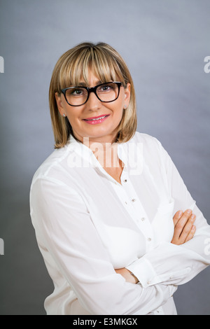 Wissenschaftliche attraktiven Frau mittleren Alters blond in Gläsern Stand mit verschränkten Armen gegen eine graue Zeitmessung in die Kamera schaut Stockfoto