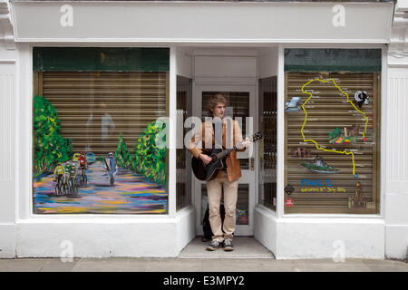 Straßenmusiker vor einem leeren Laden mit bemalten Fenstern in Skipton, Yorkshire Dales National Park, Großbritannien. Juni 2014. Alexander Johnston-Seymore Musiker & Window Art während Yorkshire sich auf die Tour de France vorbereitet, indem er die Route mit gelben Bikes und Bannern dekoriert, während sich die Unternehmen auf das weltgrößte Radrennen vorbereiten, das am 5th. Und 6th. Juli 2014 in der Grafschaft startet und Millionen von Fans zu sich bringt Am Straßenrand von Yorkshire, um die Champions des Sports anzufeuern. Stockfoto