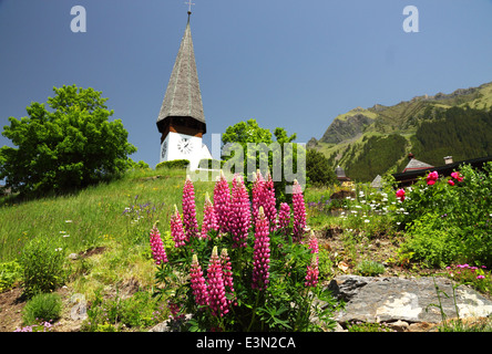 Eine weiße alpine Kirche mit einer Uhr und einem hohen Turm und Blumen. Stockfoto