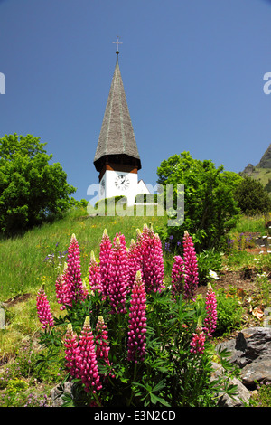 Eine weiße alpine Kirche mit einer Uhr und einem hohen Turm und Blumen. Stockfoto