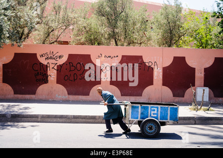 Ein Mann zieht einen Handkarren mit Waren für den Verkauf an seinem Markt entlang einer Straße Stall hinter einer Wand mit Graffiti in Englisch und Arabisch Stockfoto