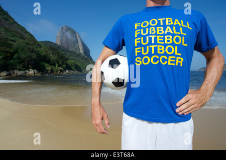 Fußballspieler im Multi Sprache Nachricht T-shirt Holding Fußball rot Strand Rio de Janeiro Brasilien Stockfoto