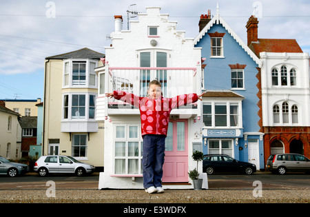 Ein junges Mädchen posiert für ein Foto mit ausgestrecktem Arm vor einem kleinen Haus an der Küste in Aldeburgh, England Stockfoto