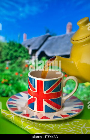 Englischer Tee Staycation Union Jack Themen Tasse, gelbe china Teekannen gießt Tee mit typischen Reetgedeckten Hütte und Garten im Hintergrund Großbritannien Stockfoto