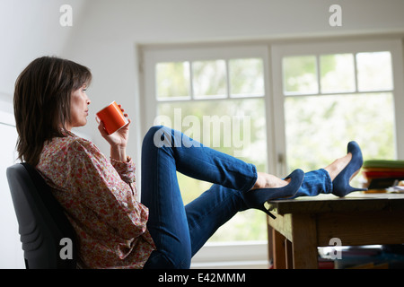 Reife Frau mit Füßen auf Schreibtisch, Kaffee trinken Stockfoto