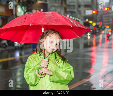 Junges Mädchen im Regen trägt einen grünen Regenmantel und hält einen roten Regenschirm von Stadt Straße Stockfoto