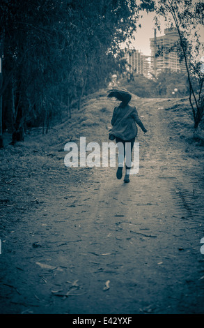Junge Frau läuft vor der Gefahr, Feldweg in Richtung Mehrfamilienhäuser Stockfoto