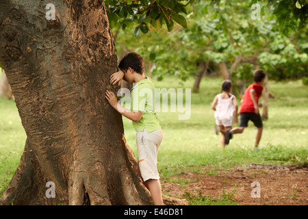 jungen und Mädchen spielen hide and seek im Park, mit Kind zählen stützte sich auf Baum Stockfoto