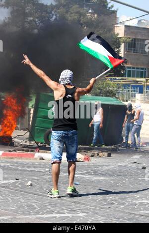 Eine palästinensische Jugend hält trotzig eine palästinensische Fahne und Wellen-Peace-Zeichen in Richtung der israelischen Streitkräfte auf der anderen Seite der Straße verbarrikadiert. Zusammenstöße brach zwischen palästinensischen Jugendlichen und Offiziere der israelischen Armee nach dem Mord an 16-j hrige Muhammad Abu Khedeir. Der junge Körper wurde von der israelischen Polizei zur Autopsie genommen und dann die Israelis verweigert, geben dem Körper zurück zu den Eltern, bis sie unterzeichnen eine Erklärung abgegeben hat, dass die Tötung war keine Revanche für die drei israelischen Jugendlichen vor einer Woche angeblich von palästinensischen militanten getötet. (Foto von Anna Ferensowicz / Pacific Press) Stockfoto