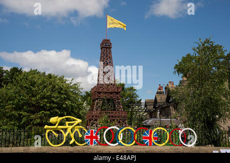 Unter blauem Himmel, Holz- Eiffelturm mit Fahne & gelb Modell der Radfahrer festen Zaun zum Garten, erbaut Le Tour in Yorkshire - Burley-In - Wharfedale, England, UK zu feiern. Stockfoto