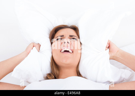 junge hübsche lässig schreienden Frau im Bett für Ohren mit Kissen wegen Lärm. Lustiges Bild. Lärm oder Schlaflosigkeit-Konzept Stockfoto