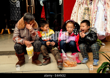 Pengzhou, China: Chinesische Seniorin sitzt auf einem Schritt vor einem Bekleidungsgeschäft mit drei glückliche Kinder Stockfoto