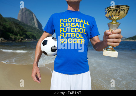 Fußballspieler in Multi Sprache Nachricht T-shirt Holding Trophy und Fußball ball rot Strand Rio de Janeiro Brasilien Stockfoto
