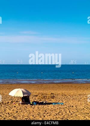 Person im Liegestuhl unter einem Sonnenschirm am Strand in Colwyn Bay North Wales Blick in Richtung Offshore-Windpark in der irischen See Stockfoto