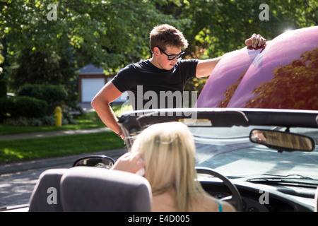 Ein junger Mann mit der Motorhaube, die Autos Probleme zu betrachten, während eine junge Frau in dem Fahrersitz tätigen eines Anrufs auf ihr sitzt Stockfoto