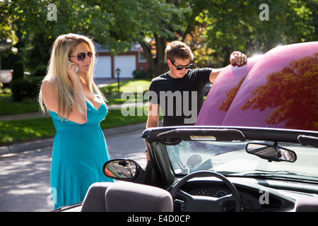 Eine junge Frauen steht neben einem aufgeschlüsselt Auto fordert einen Abschleppwagen LKW oder Auto Club Hilfe als junger Mann Kontrollen unter der Haube Stockfoto