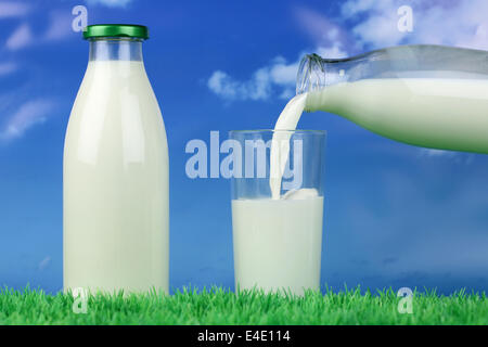 Frische Milch aus der Flasche in ein Glas gießen, auf einer Wiese vor einem blauen Himmel Stockfoto