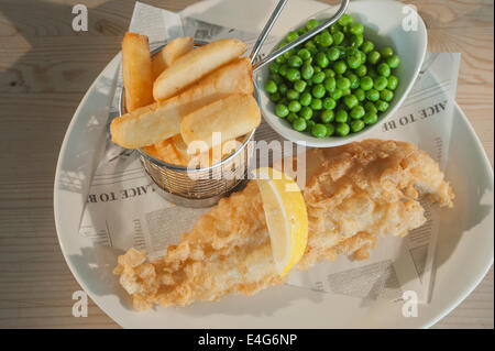 Fish &amp; Chips (frittierte Schellfisch in Teig) mit Erbsen und ein Segment der Zitrone auf einem weißen Teller auf einem Holztisch Stockfoto