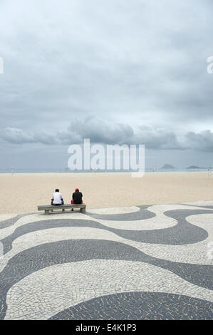 Figuren sitzen auf Bank am berühmten Bürgersteig Fliesenmuster vor leeren Blick auf Copacabana Strand Rio de Janeiro Brasilien unter grauem Himmel Stockfoto