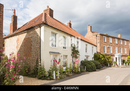 Hübsche weiße Häuschen mit im Vorgarten wachsenden Hollyhocks und einem großen Stadthaus in Burnham Market, einer kleinen Stadt im Norden Norfolks, Blick auf die Straße Stockfoto