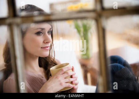 Junge Frau, die Betrachtung im Wohnzimmer mit Kaffeetasse in Händen Stockfoto