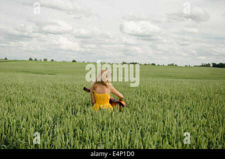 Blondes Mädchen mit einem gelben Sommerkleid sitzt im Bereich der Roggen spielt die Gitarre und swing Stockfoto