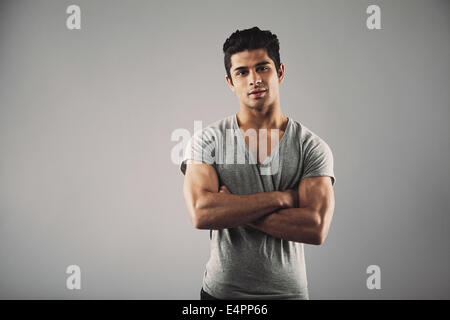 Porträt des jungen muskulöser Mann posiert vor grauem Hintergrund. Hübscher Junge hispanic Männermodel stehend mit seinen Armen gekreuzt