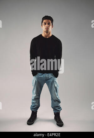 In voller Länge Portrait von stilvollen junger Mann in Casuals überragt mit seinen Händen in Tasche grau hinterlegt. Stockfoto