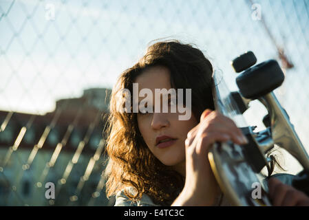 Close-up Portrait von Teenager-Mädchen stehen im Freien neben Maschendrahtzaun, halten Skateboard und Blick in die Kamera, Deutschland Stockfoto