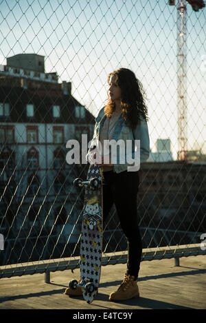 Teenager-Mädchen stehen im Freien neben Maschendrahtzaun in der Nähe von Comercial Dock mit Skateboard, Deutschland Stockfoto