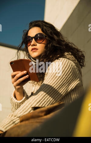 Close-up Portrait von Teenager-Mädchen im Freien mit Sonnenbrille, sitzt neben Aufbau und halten Smartphone, Deutschland Stockfoto