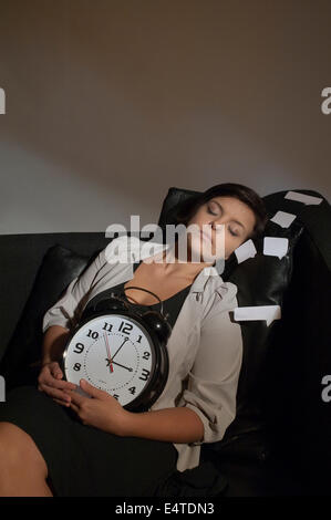 Porträt von Mid-Adult Woman schlafen auf Sofa und hält Wecker umgeben von Bubble Klebstoff Notizen Stockfoto