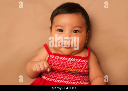 Close-up Portrait von asiatisches Baby auf Rücken liegend, roten Kleid, Blick in die Kamera und machen lustige Gesicht, Studio gedreht Stockfoto