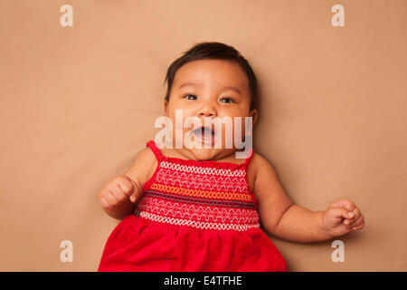 Close-up Portrait asiatisches Baby auf Rücken liegend, roten Kleid, Blick in die Kamera und lächelnd, erschossen Studio auf braunem Hintergrund Stockfoto