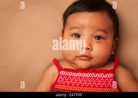 Close-up Portrait von asiatisches Baby auf Rücken liegend, roten Kleid, Blick in die Kamera, Studio gedreht auf braunem Hintergrund Stockfoto