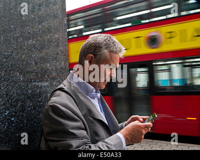 Reife Geschäftsmann mit ein iPhone Smartphone in London Southbank mit unscharfen traditionelle London red Bus vorbei im Hintergrund Stockfoto