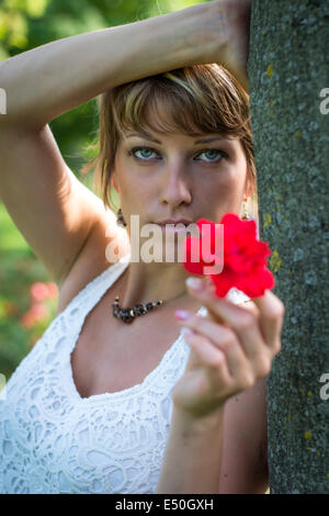 Attraktive Frau in eine stilvolle weiße Spitzen oben streckte eine duftende rote rose symbolisch für Liebe, wie sie lehnt sich gegen einen Baum in Stockfoto