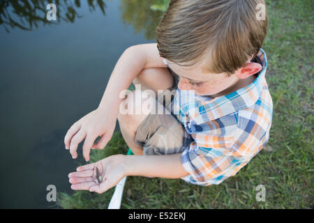 Ein kleiner Junge im Freien sitzen am Ufer eines Flusses mit einem kleinen Fisch in seiner Handfläche. Stockfoto