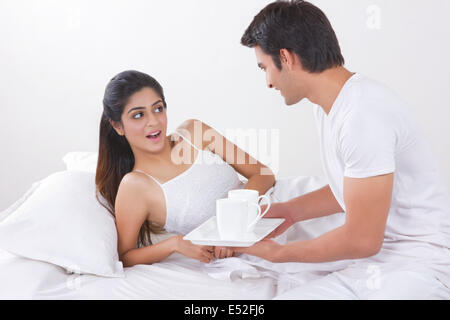 Junger Mann hält Tablett mit Kaffeetassen von überrascht Frau im Bett Stockfoto