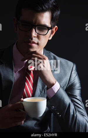 Nachdenklicher junger Geschäftsmann mit Kaffeetasse vor schwarzem Hintergrund Stockfoto