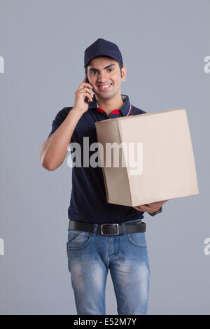 Porträt der Lieferung Mann trägt Paket während der Beantwortung Handy vor grauem Hintergrund Stockfoto