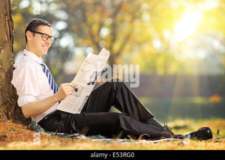 Junger Mann mit Krawatte sitzt auf einer Wiese und liest eine Zeitung in einem park Stockfoto